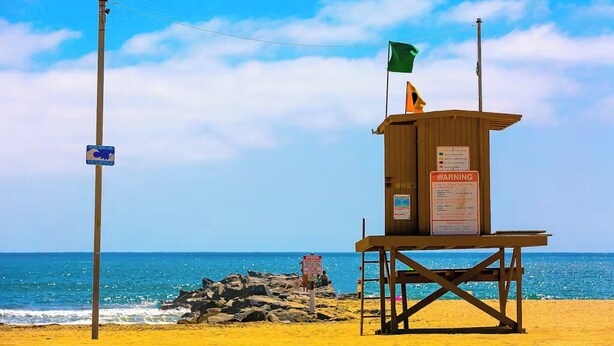 A lifeguard tower on a beach with a flag, a warning sign, and a blue sign on a pole. - Cozy 2-Bedroom Getaway (Newport Beach)