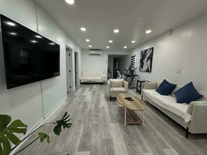 A modern living room with a flat-screen TV, a sofa with blue cushions, a coffee table, and a plant. - Updated and Comfortable Trailer in West OC! (Berlin)