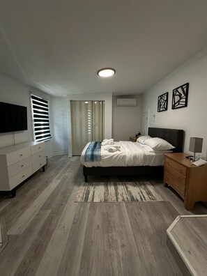 A bedroom with a bed, bedside tables, a TV, a wardrobe, a window with blinds, and an air conditioning unit. - Updated and Comfortable Trailer in West OC! (Berlin)