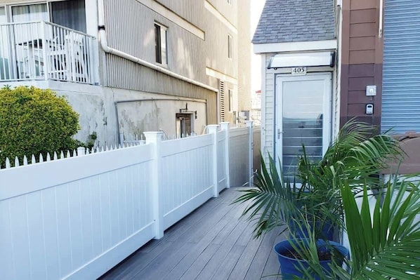 A narrow wooden deck with a white fence, a potted plant, and a building with a number '405' sign.