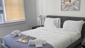 Ein ordentlich bezogenes Bett mit weißen Laken, einem grauen Kopfteil und einer blauen, strukturierten Decke.