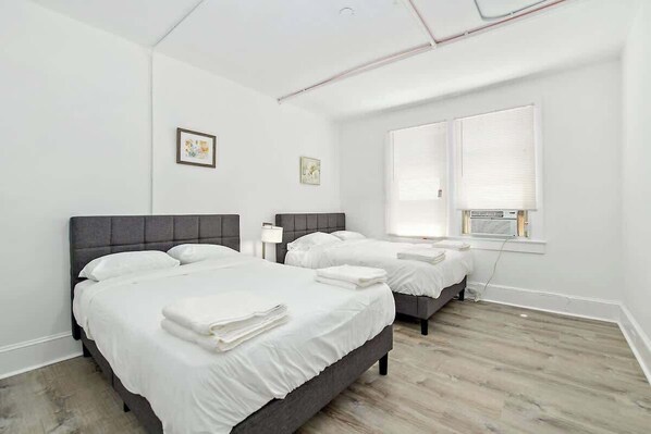 Un dormitorio con dos camas, ropa de cama blanca, un ventanal con persianas y un techo con tuberías a la vista.