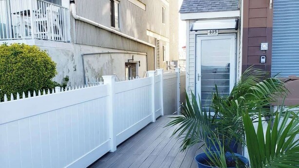 Una terraza de madera con una cerca blanca, una planta en maceta y un edificio con una puerta cerrada marcada con el número 405.