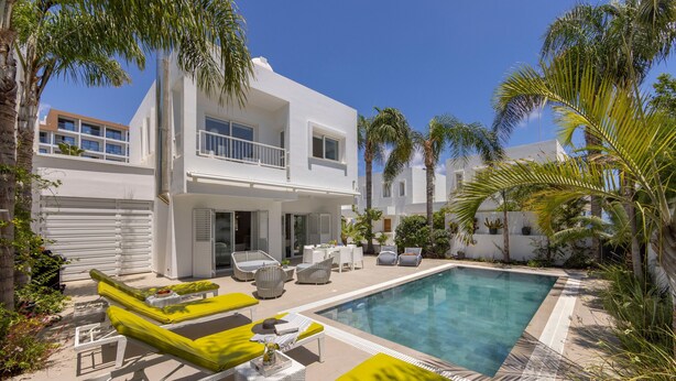 Une maison moderne à deux étages, dotée d’une piscine, de mobilier d’extérieur et de palmiers.