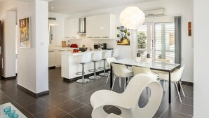 Une cuisine moderne avec des meubles blancs, un îlot central et des tabourets de bar. On y trouve également un coin repas avec une table en verre et des chaises blanches.