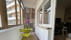A narrow balcony with a small table and chairs, a potted plant, and a view of a building with multiple floors.