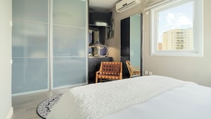 Ein Schlafzimmer mit einem Bett, einem Holzstuhl, einem Kleiderschrank und einem Fenster mit Blick auf Gebäude.