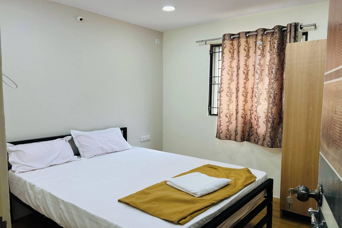 A single bed with white bedding, a brown throw blanket, and a window with patterned curtains.