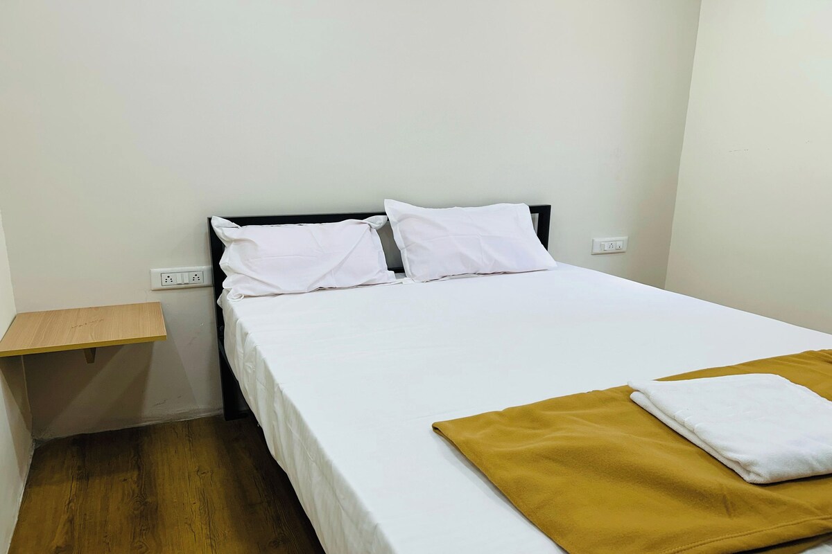 A single bed with white bedding and a mustard-colored blanket, a wooden nightstand with a power outlet, and a plain white wall.