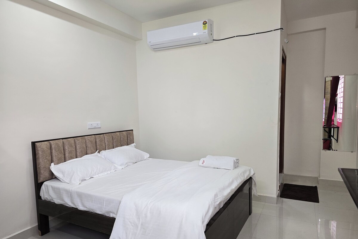 A neatly made bed with white bedding in a room with white walls and a ceiling-mounted air conditioner.