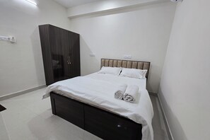 Un lit bien fait, avec du linge de lit blanc et des serviettes pliées, une armoire en bois foncé et un sol carrelé blanc et impeccable dans une chambre d’hôtel.