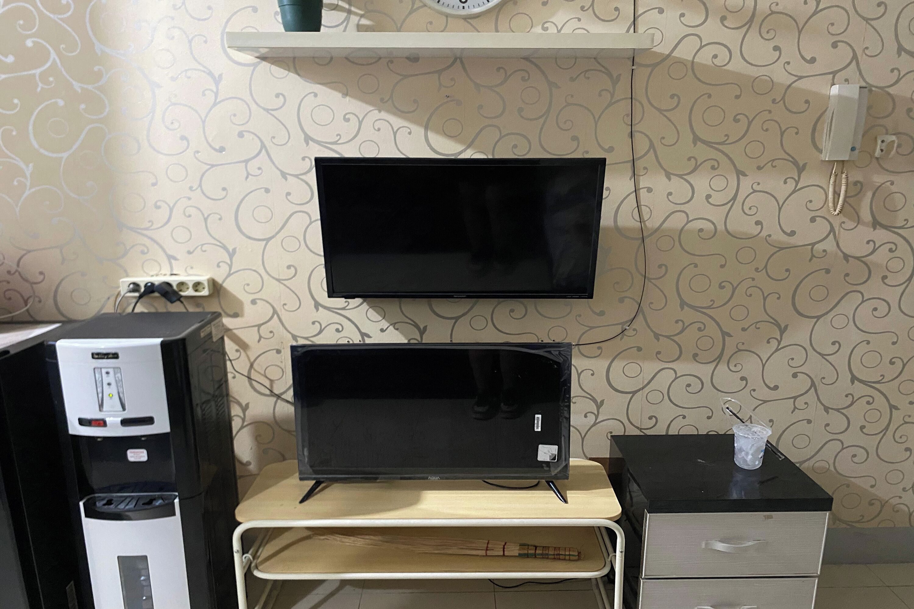 A room with a flat-screen TV mounted on the wall, a desktop computer, a water dispenser, and a small table with a broom.