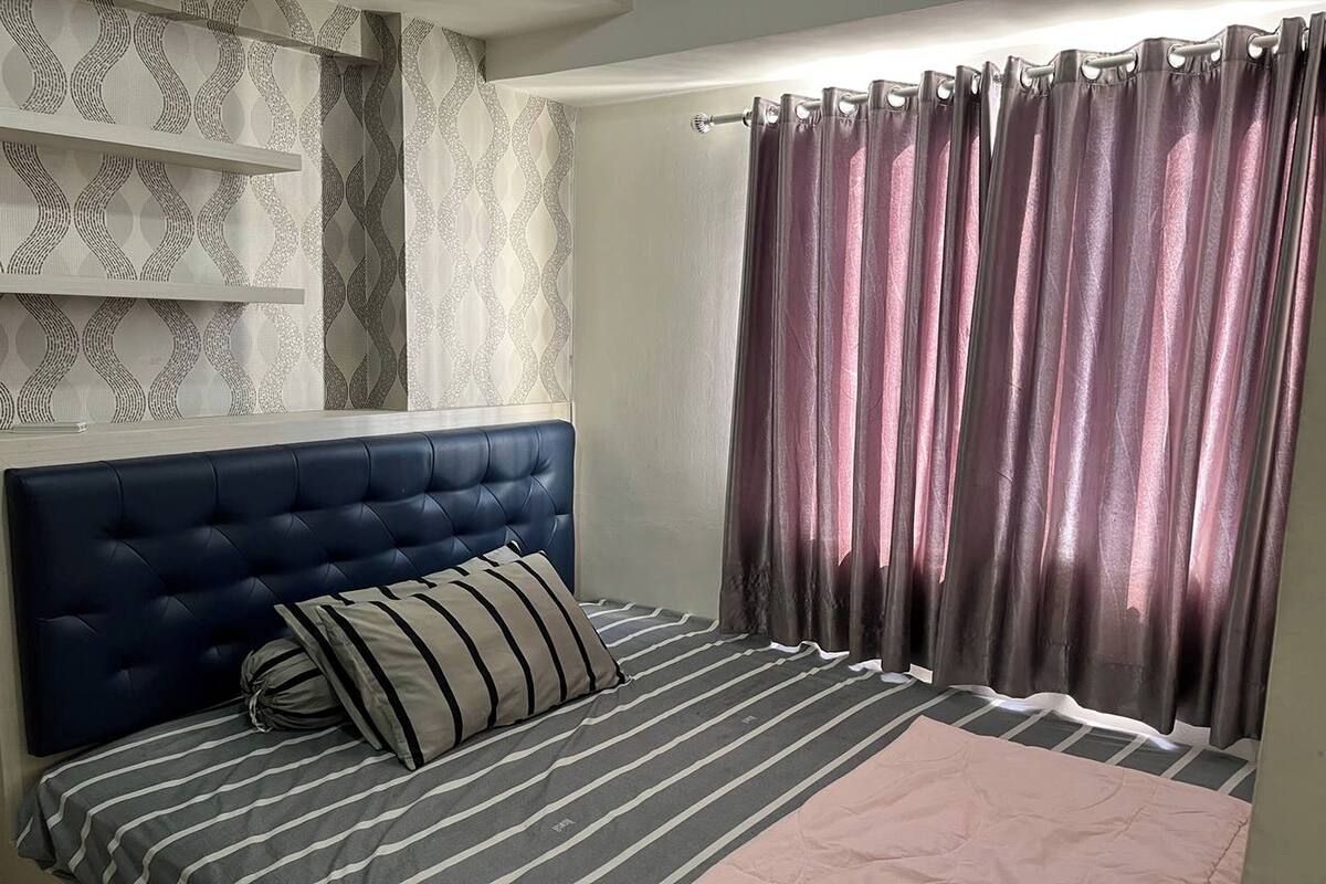 A bedroom with a tufted headboard, striped bedding, a pink throw, and a window with dark curtains.