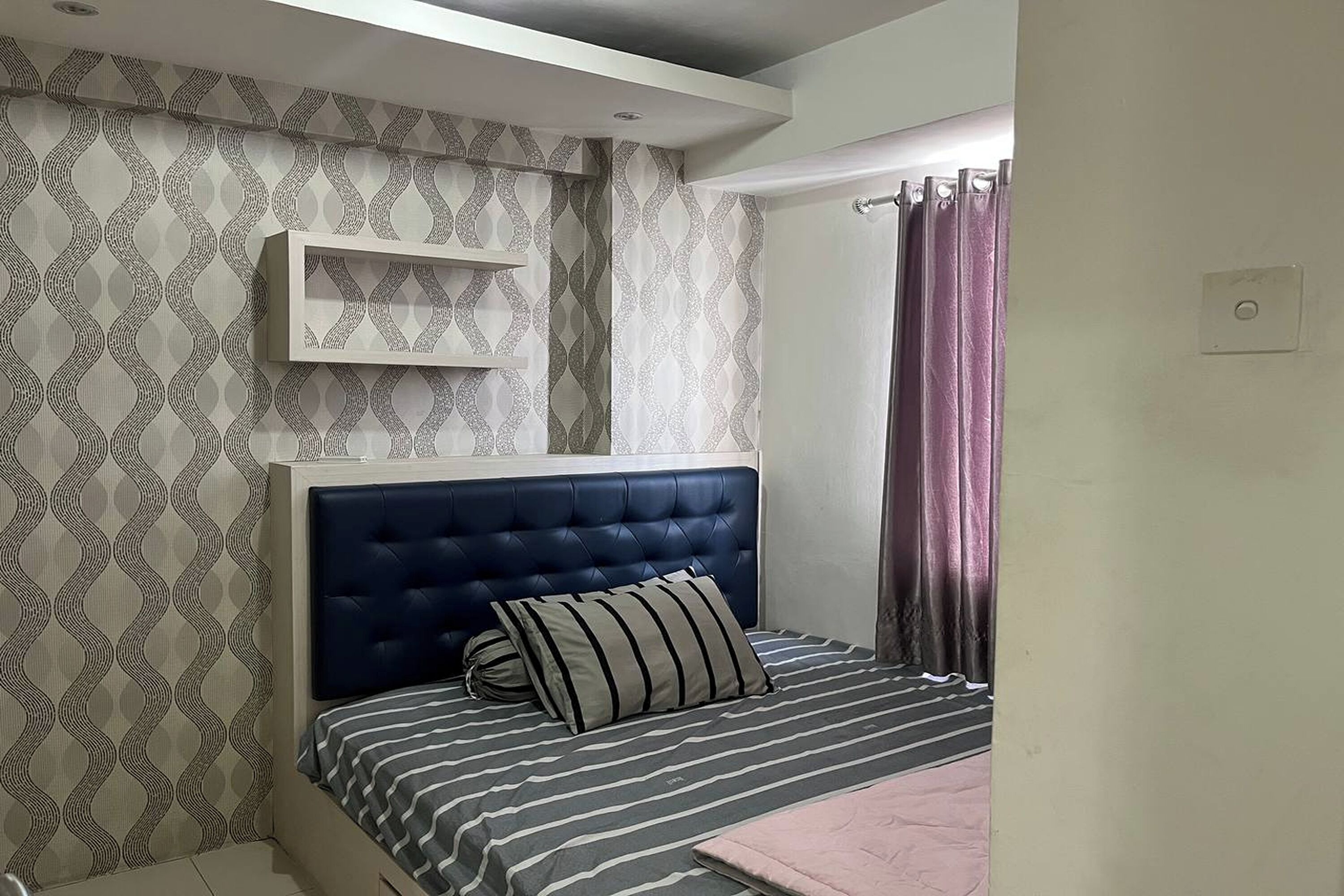Une chambre à coucher avec une tête de lit capitonnée, une couvre-lit à rayures et un papier peint à motifs.