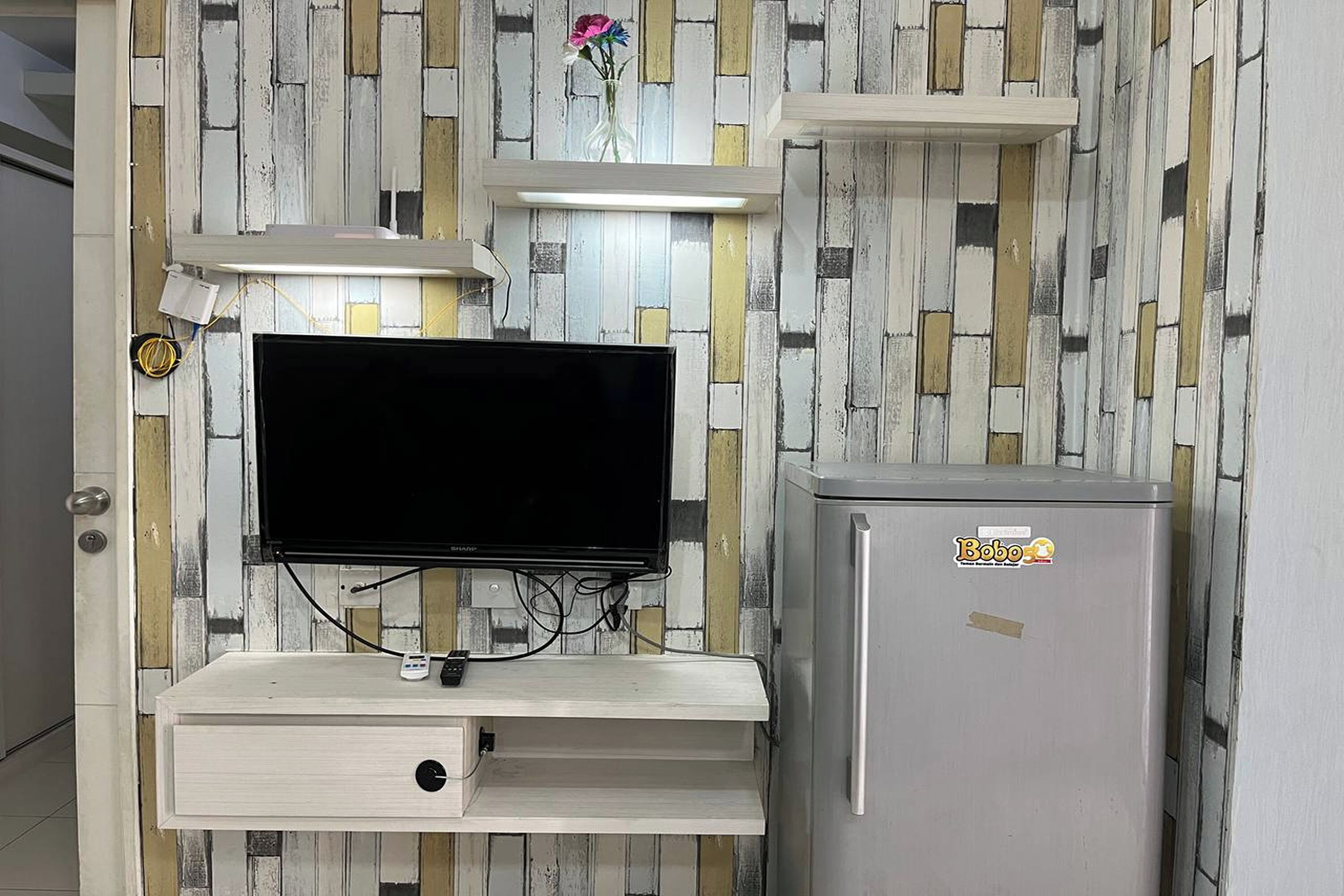 Ein Zimmer mit einem an einer Holzablage montierten Flachbildfernseher, einem kleinen Kühlschrank mit Aufkleber und einer Wand mit farbigem Holzdekor.