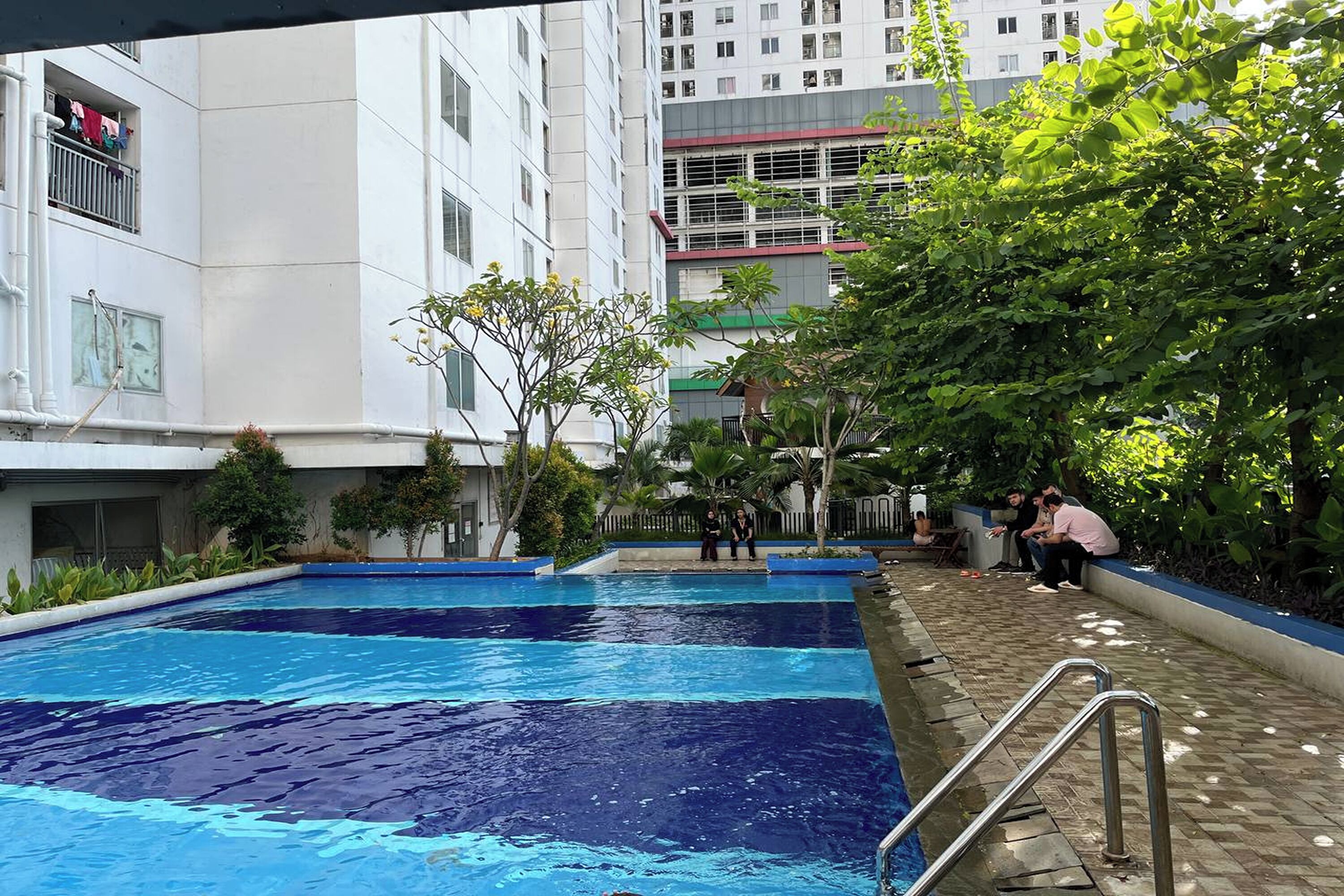 Una piscina con solárium de baldosas, rodeada de vegetación y edificios.