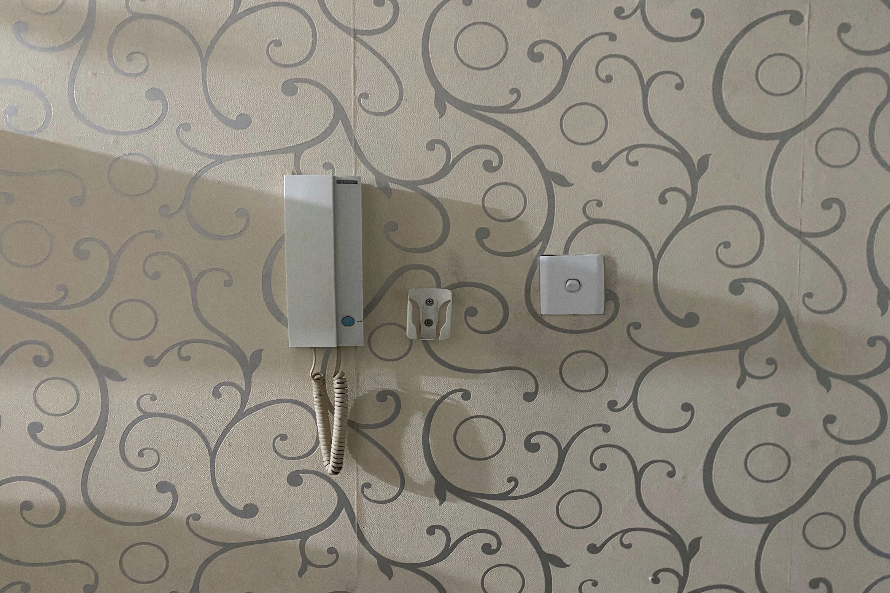Un téléphone fixé au mur, doté d’un fil et d’un petit bouton, sur un fond de papier peint à motifs.