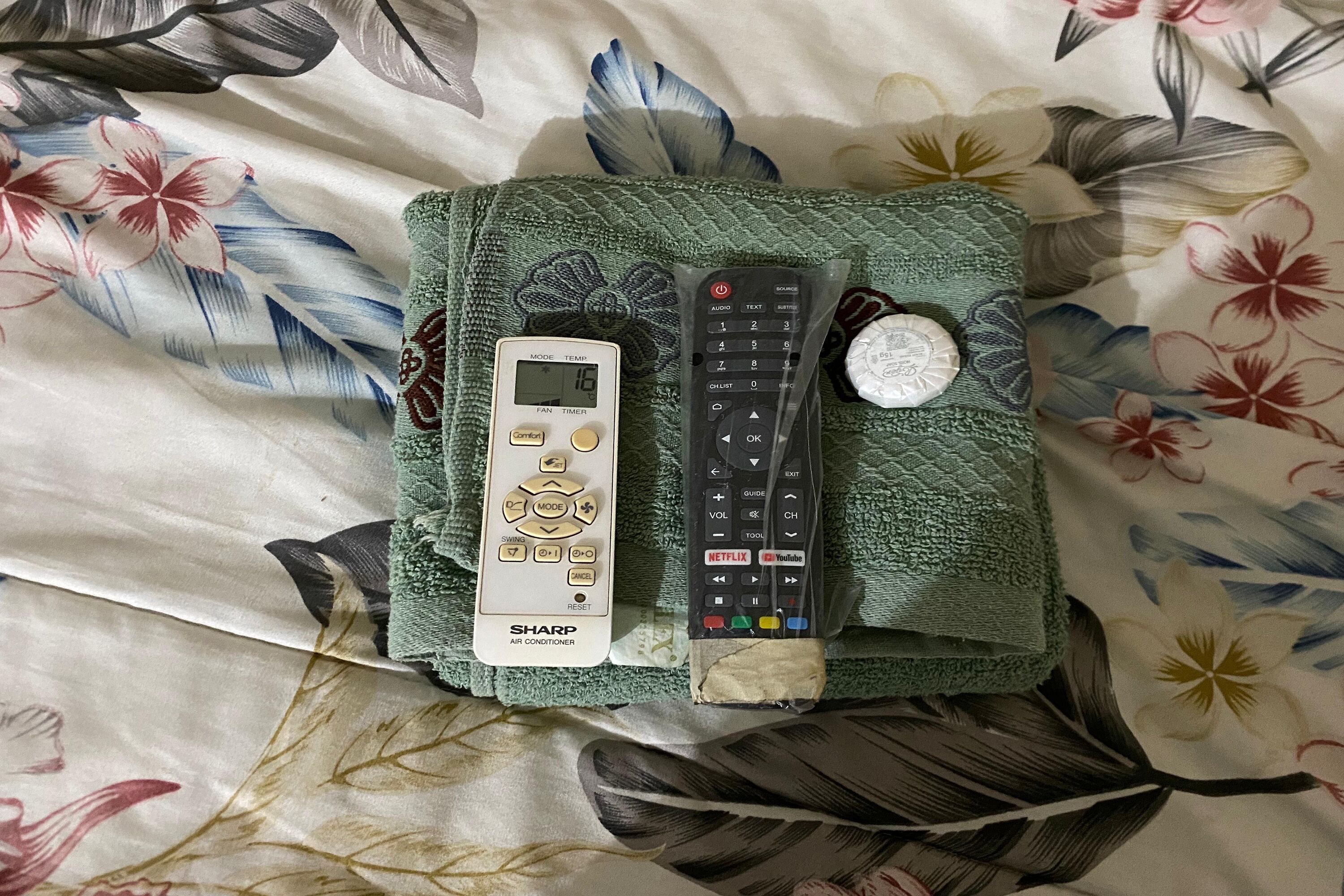 A bed with a floral-patterned bedspread, a Sharp air conditioner, and two remote controls.