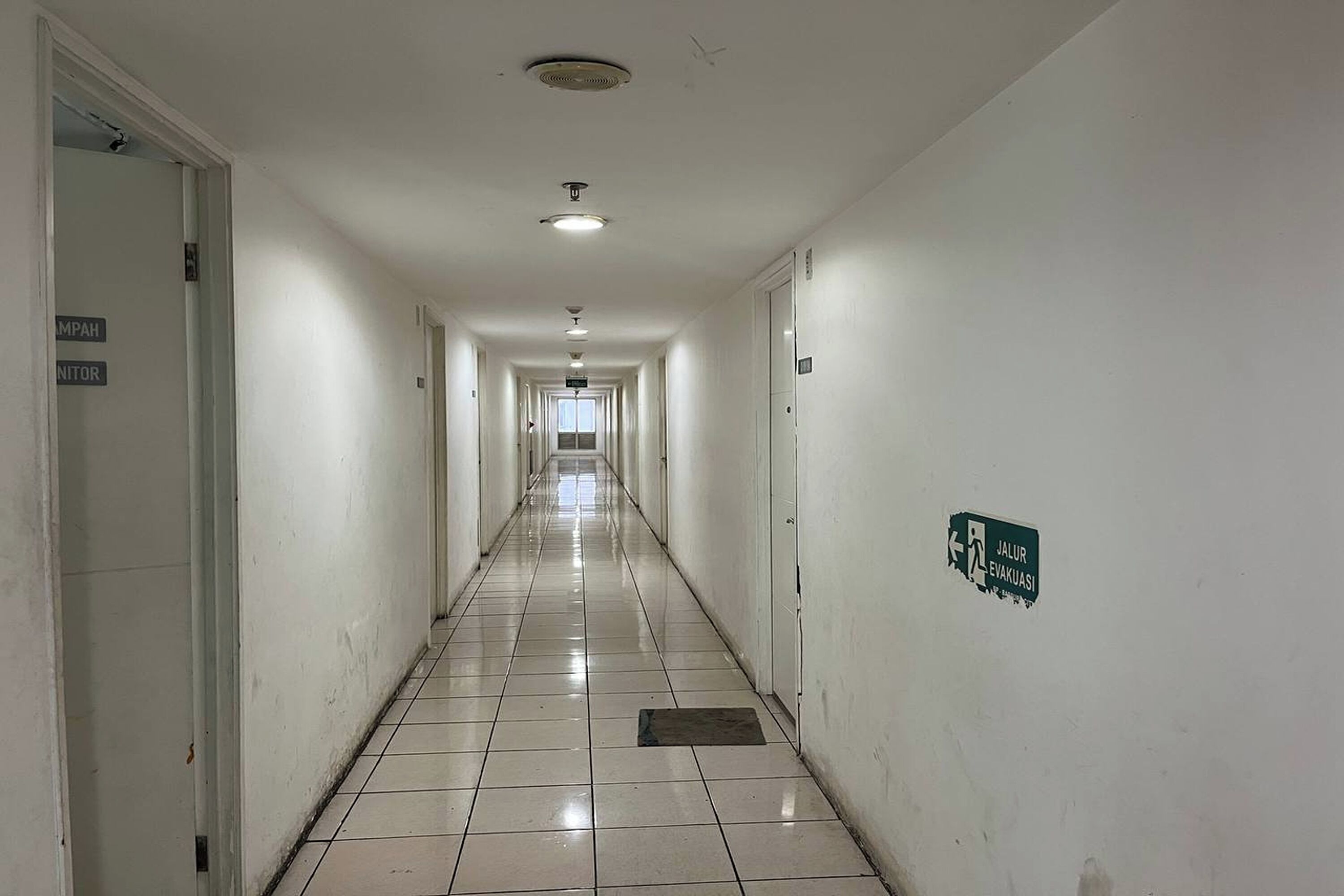 A long, narrow hallway with white walls and tiled floors. There are doors on both sides, and a green sign on the right wall indicating an evacuation route.