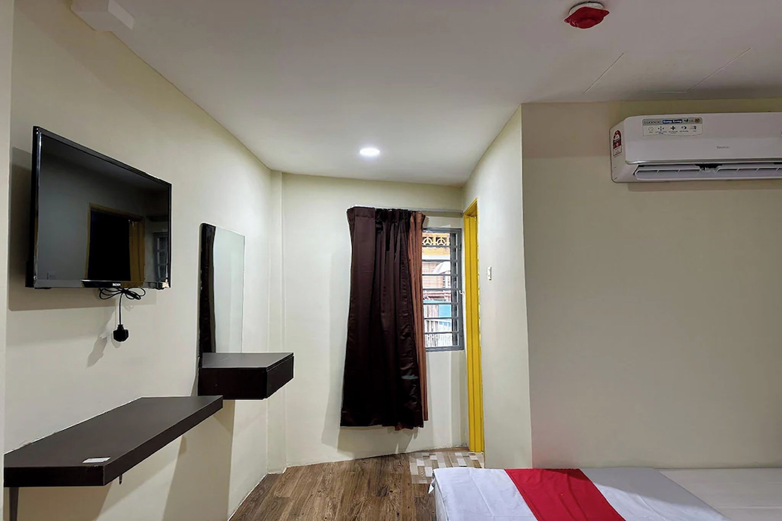 A small, clean room with a bed, a mounted TV, a wall-mounted air conditioner, and a window with curtains.
