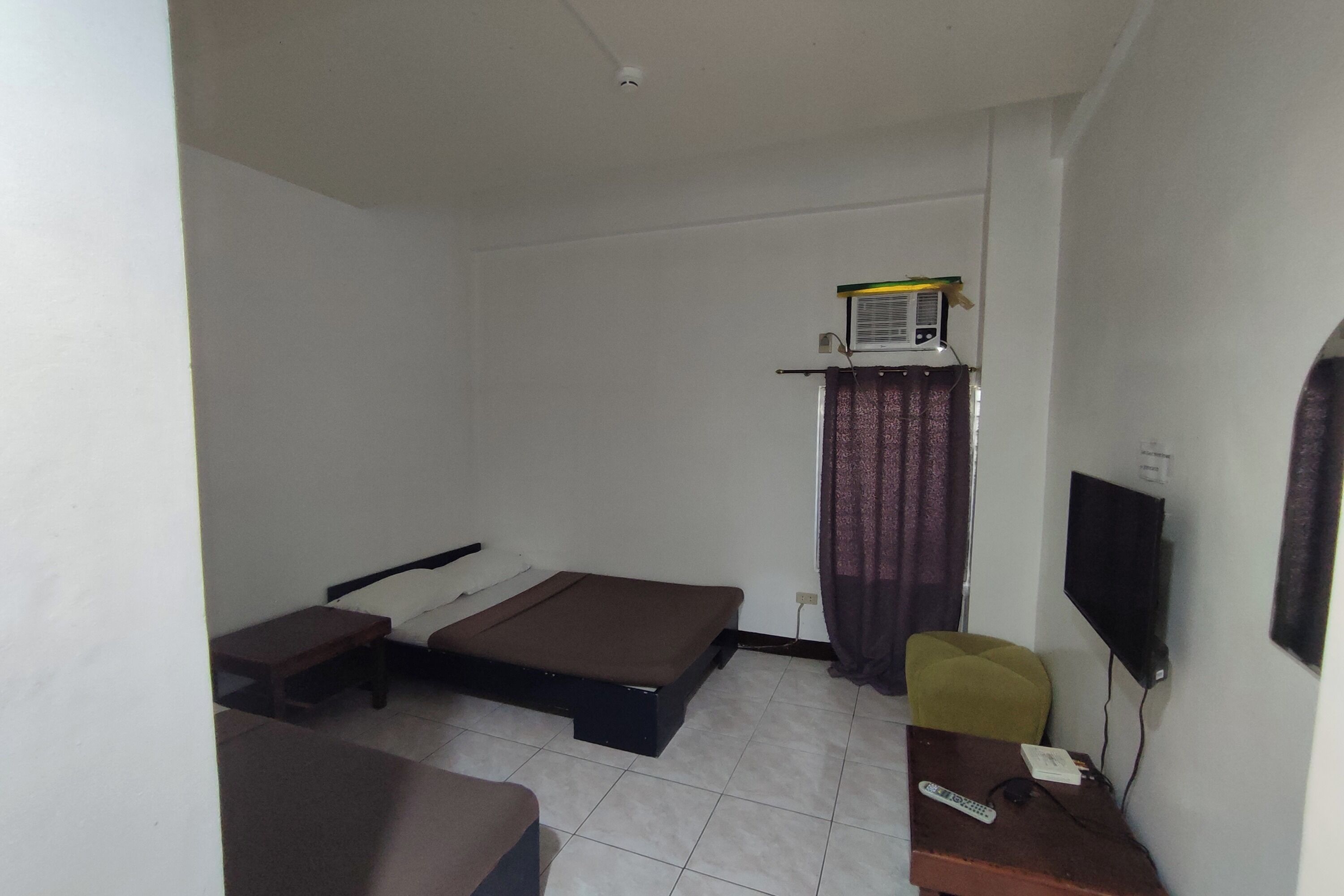 A small, sparsely furnished room with a bed, a desk, a chair, a television, and a window with curtains.