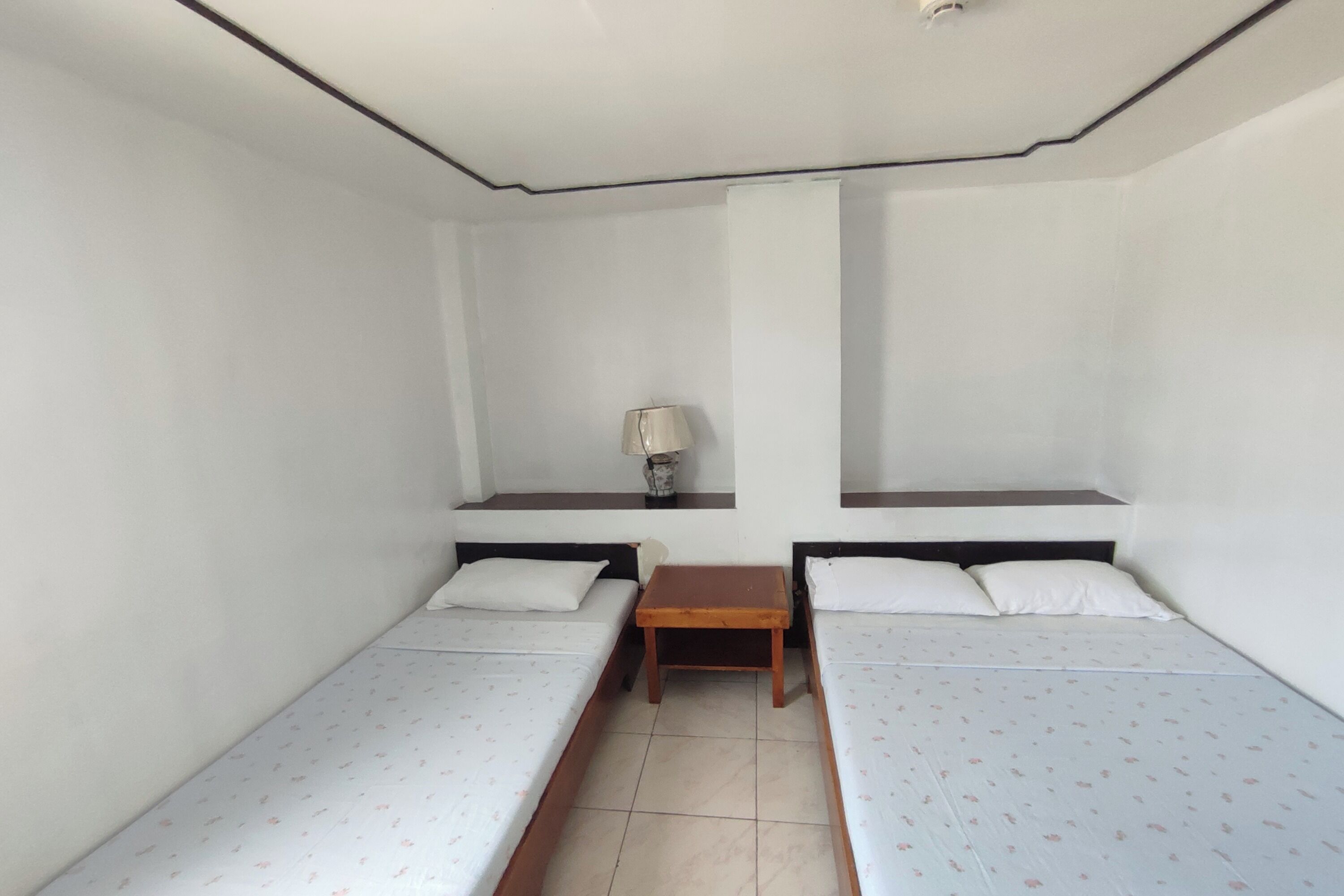 A small, clean room with two single beds, a small table, and a lamp on a shelf.