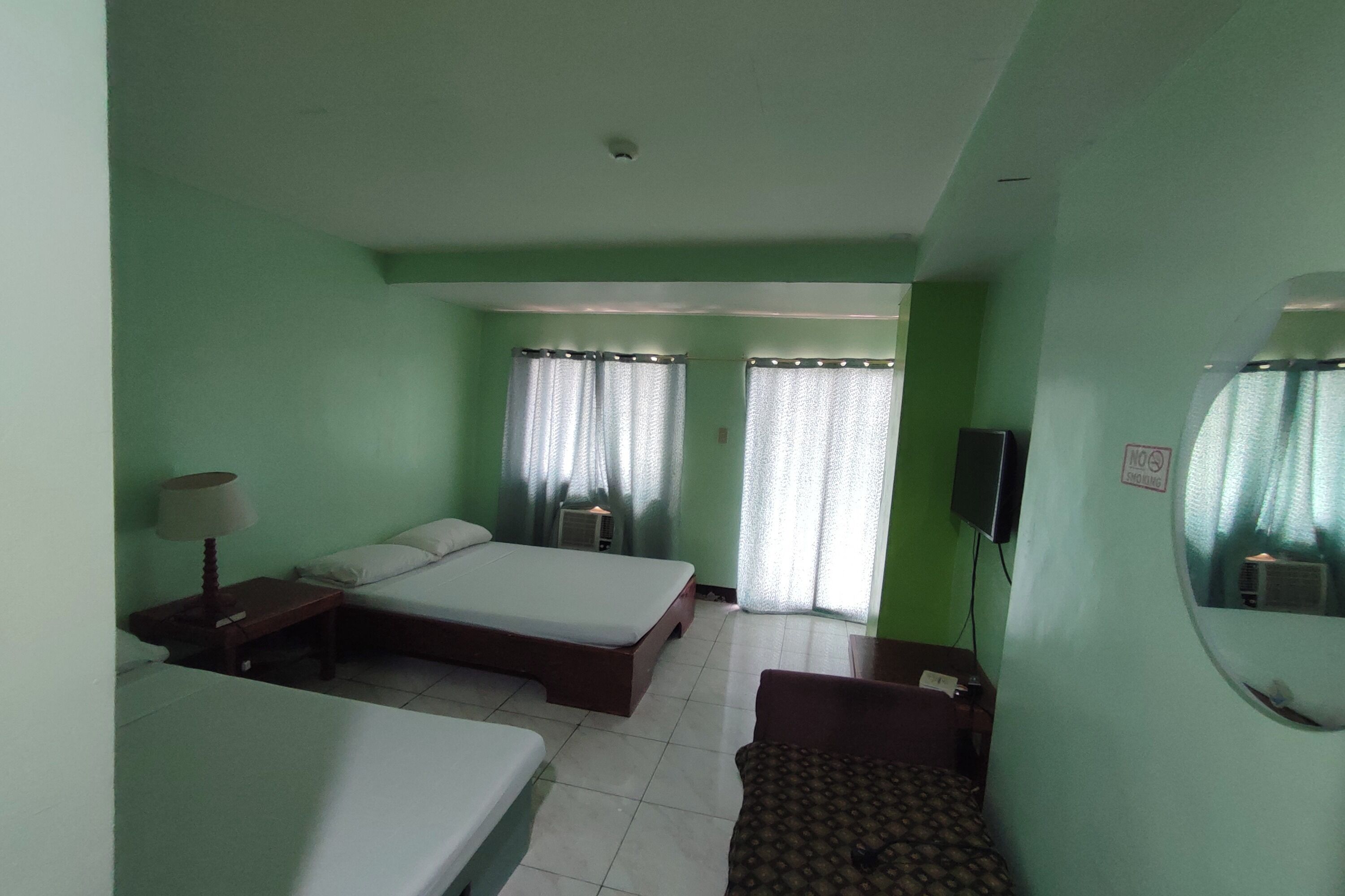 A hotel room with two single beds, a small table, a chair, a lamp, a TV, and a mirror.