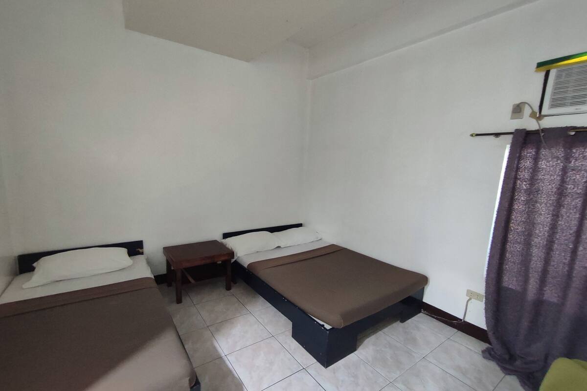 Una habitación pequeña y limpia con dos camas individuales, una mesita y una cortina en la ventana.