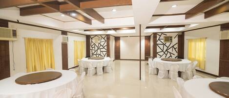 A spacious, well-lit room with multiple round tables, each surrounded by white chairs. The room features a high ceiling with exposed beams and a polished floor.