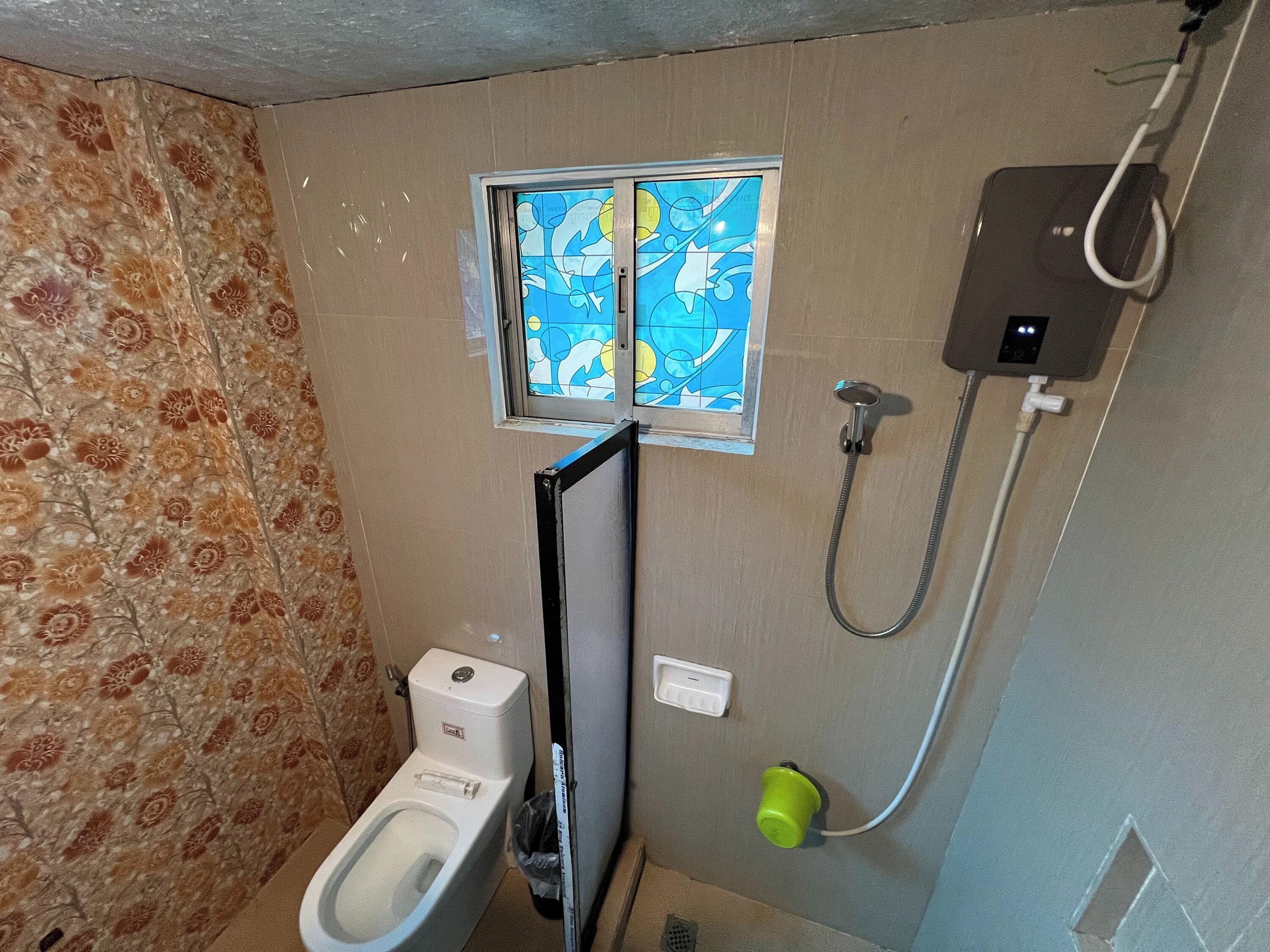 Un baño con inodoro, ducha y ventana con diseño floral.