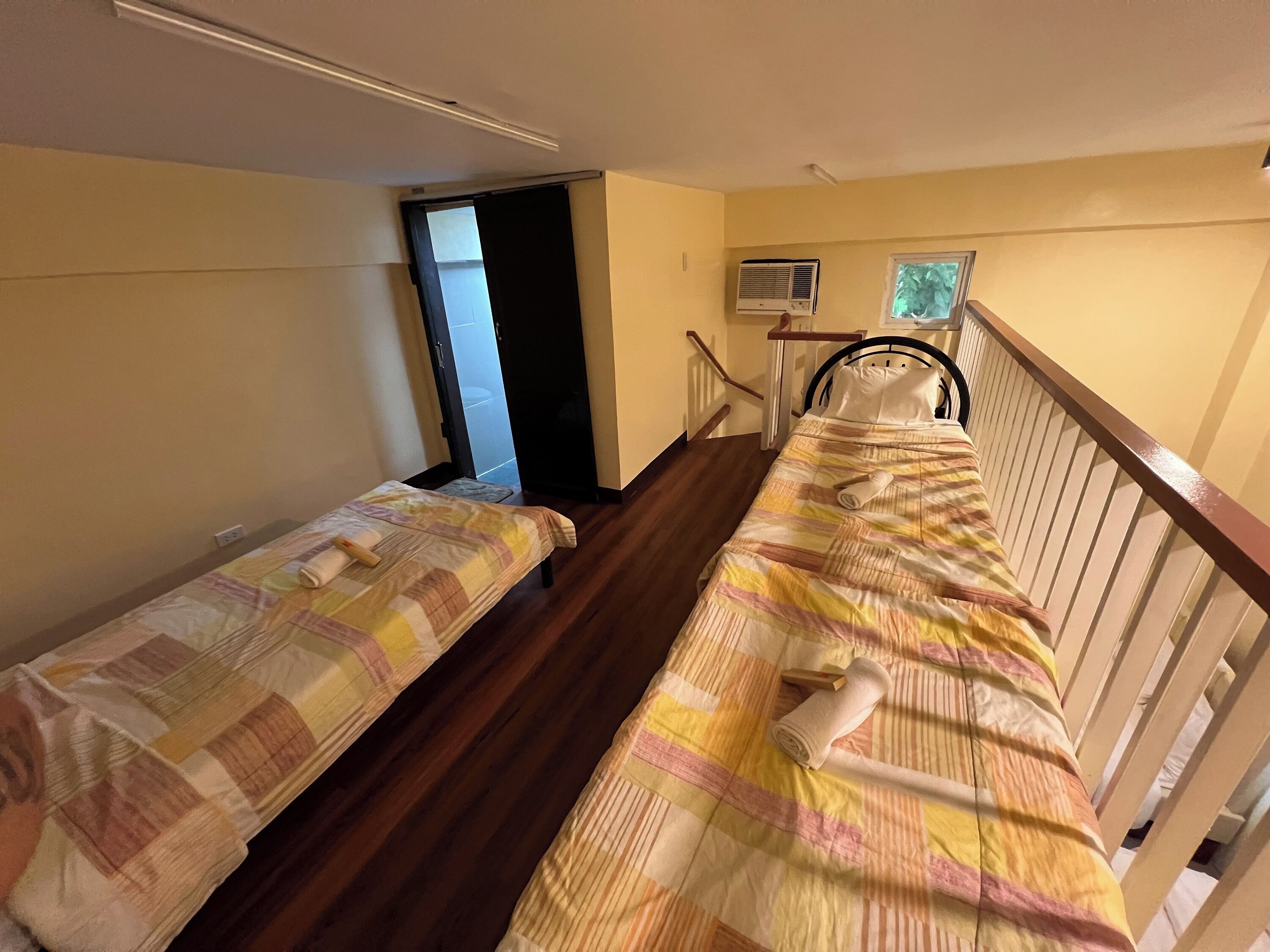 A room with two beds, wooden flooring, a window with blinds, a small air conditioning unit, and a door leading to another room.
