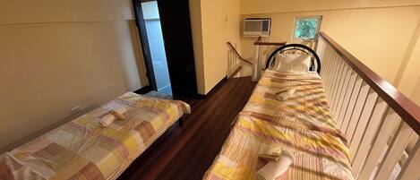 A room with two beds, wooden flooring, a window with blinds, a small air conditioning unit, and a door leading to another room.