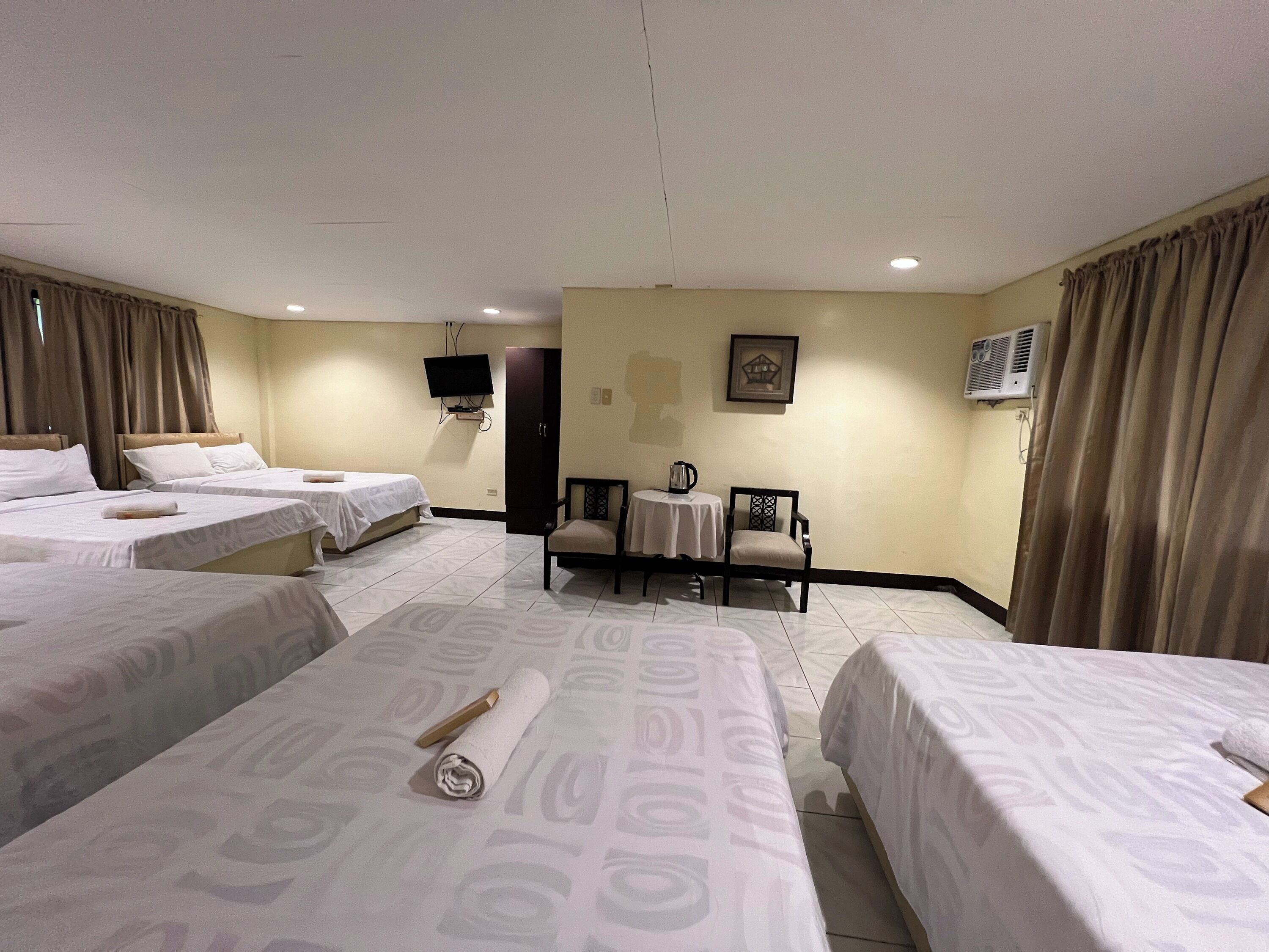 Quarto de hotel com quatro camas individuais, uma pequena mesa com chaleira, duas cadeiras, uma TV fixada na parede e um aparelho de ar condicionado.