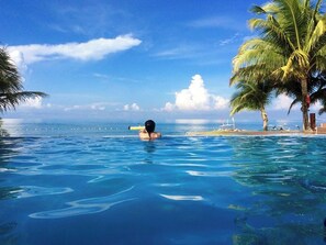 A clear blue pool with a person swimming, surrounded by palm trees and a view of the ocean. - Vista Venice Resort (Morong)