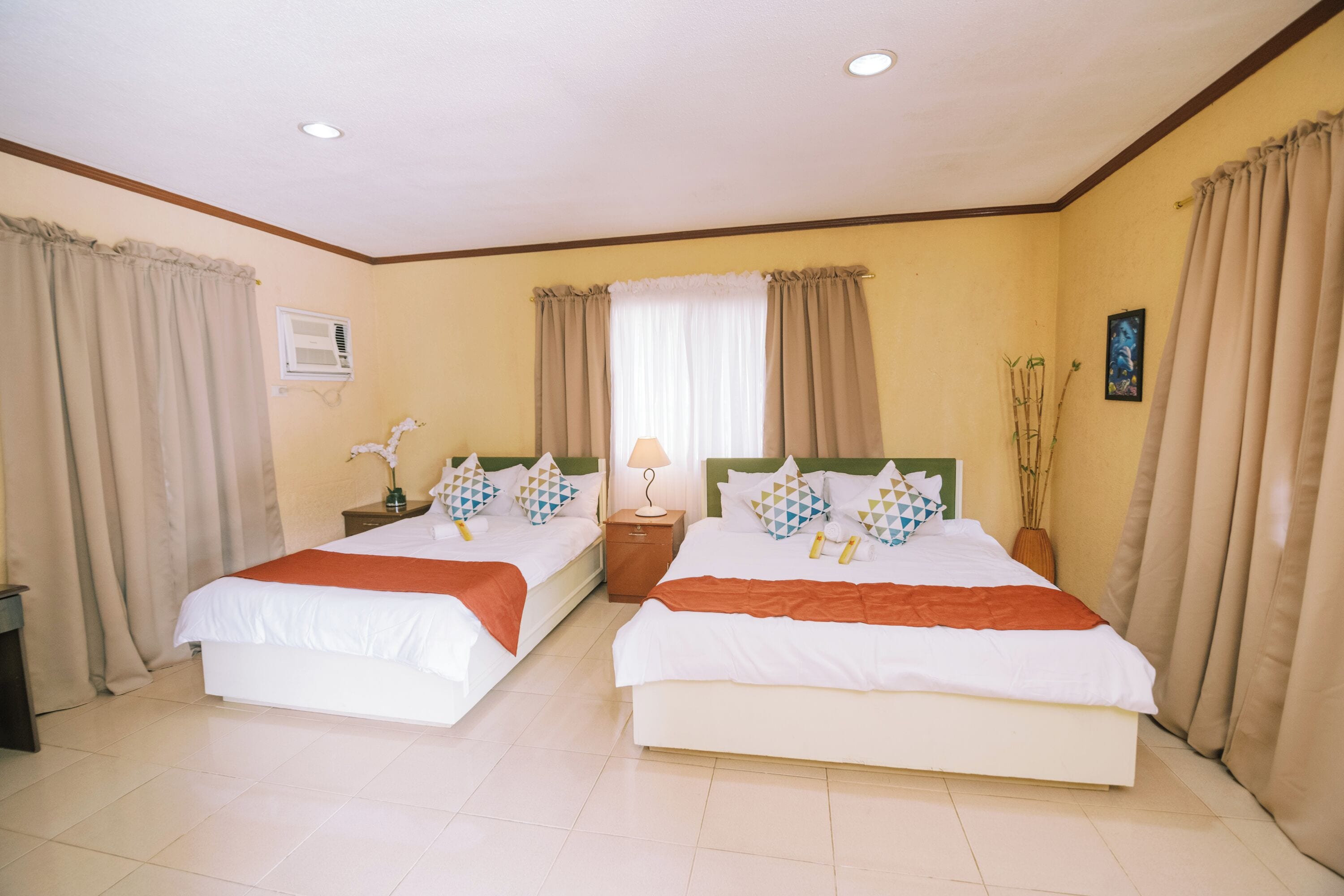 A hotel room with two beds, a window with curtains, a bedside table with a lamp, and a wall-mounted air conditioner.