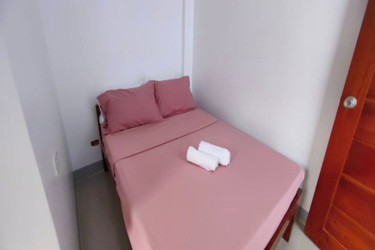 Una cama pequeña y ordenada con ropa de cama rosa y dos toallas enrolladas.