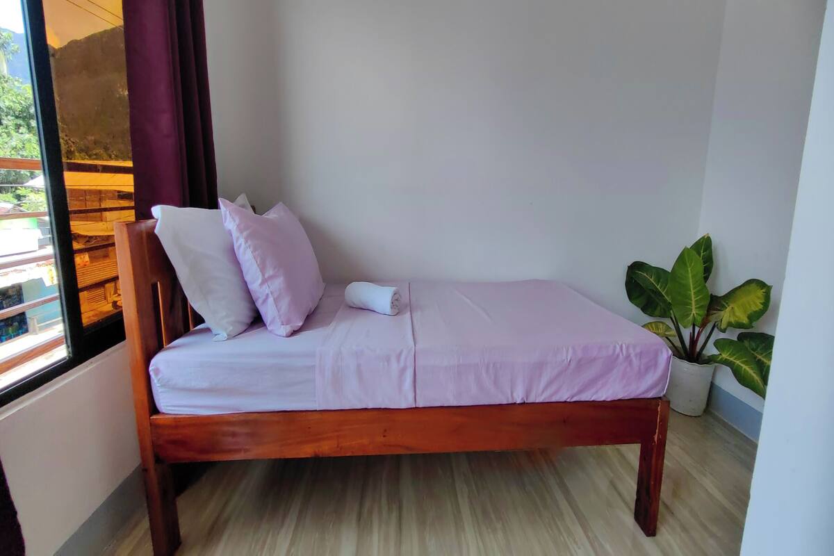Un lit simple avec un cadre en bois et un matelas de couleur claire. Une plante en pot se trouve à droite du lit.