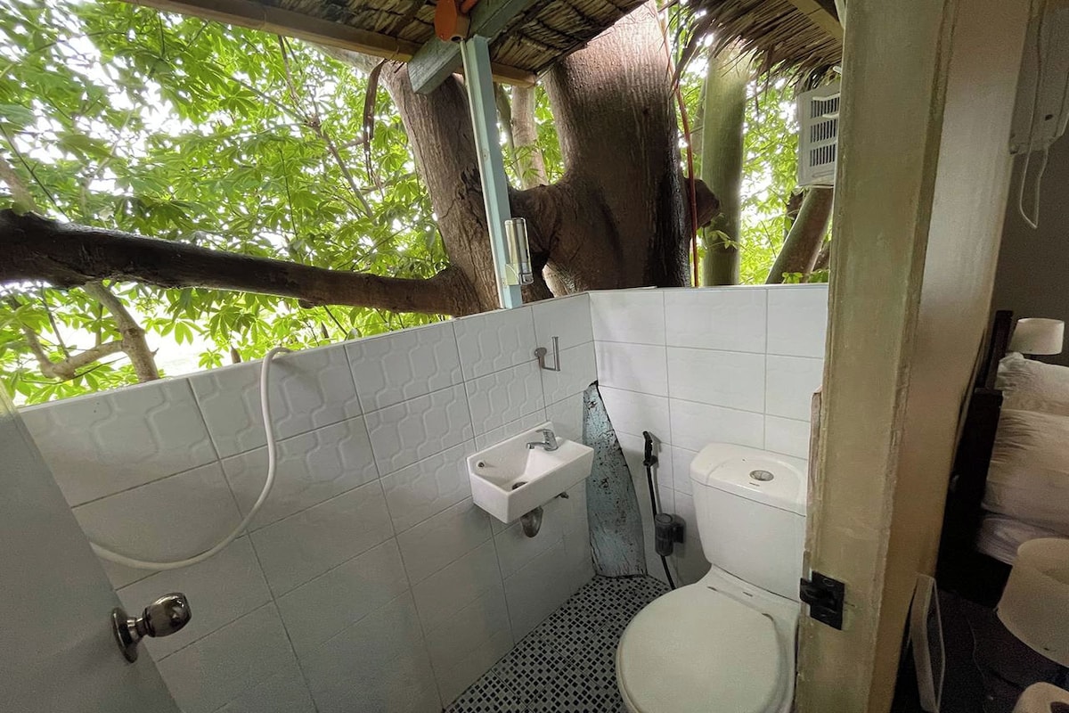 Un baño con lavamanos, inodoro y zona de ducha blancos, rodeado de paredes con azulejos y un árbol en el exterior.
