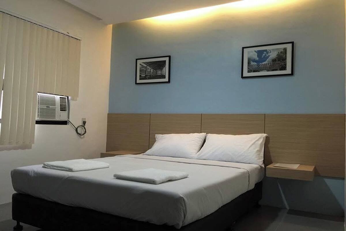 Quarto de hotel moderno com uma cama bem arrumada, uma mesinha de cabeceira e duas fotos emolduradas na parede.