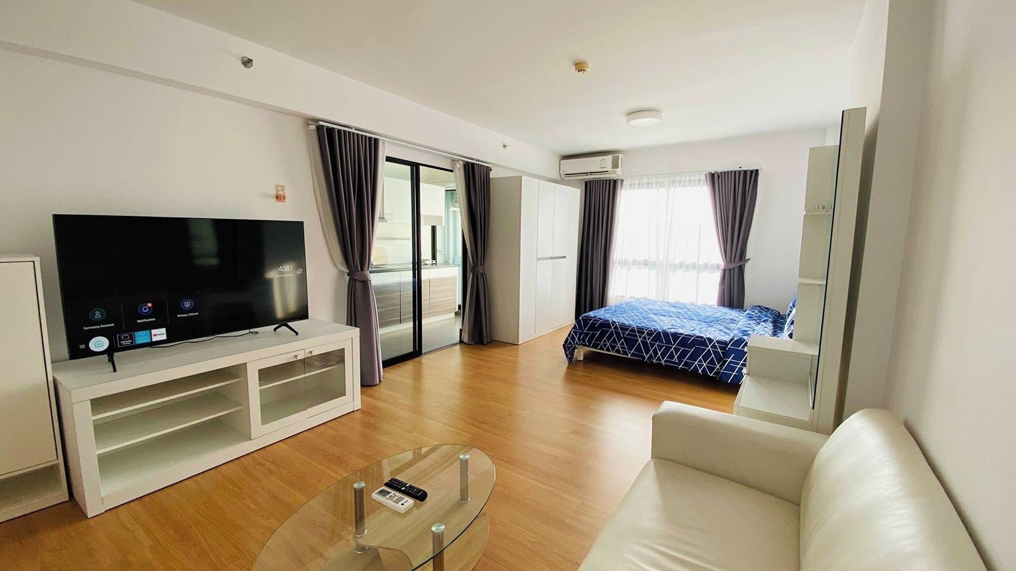 A modern hotel room with a flat-screen TV, a glass coffee table, a sofa, a bed with a blue patterned bedspread, and a sliding door leading to a balcony.