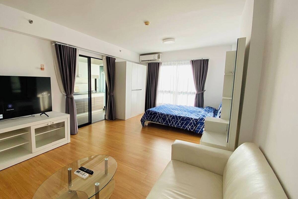 Une chambre d’hôtel moderne équipée d’un téléviseur à écran plat, d’une table basse en verre, d’un canapé, d’un lit recouvert d’une courtepointe à motifs bleus et d’une porte coulissante donnant sur un balcon.