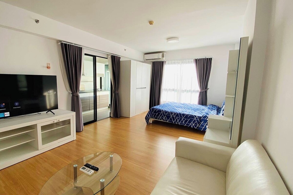 A modern hotel room with a flat-screen TV, a glass coffee table, a sofa, a bed with a blue patterned bedspread, and a sliding door leading to a balcony.