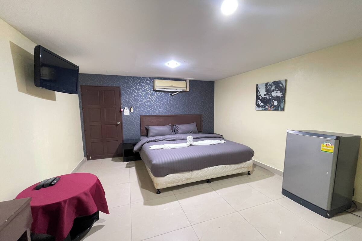 A hotel room with a bed, a television mounted on the wall, an air conditioner, a refrigerator, and a small table with a red tablecloth.