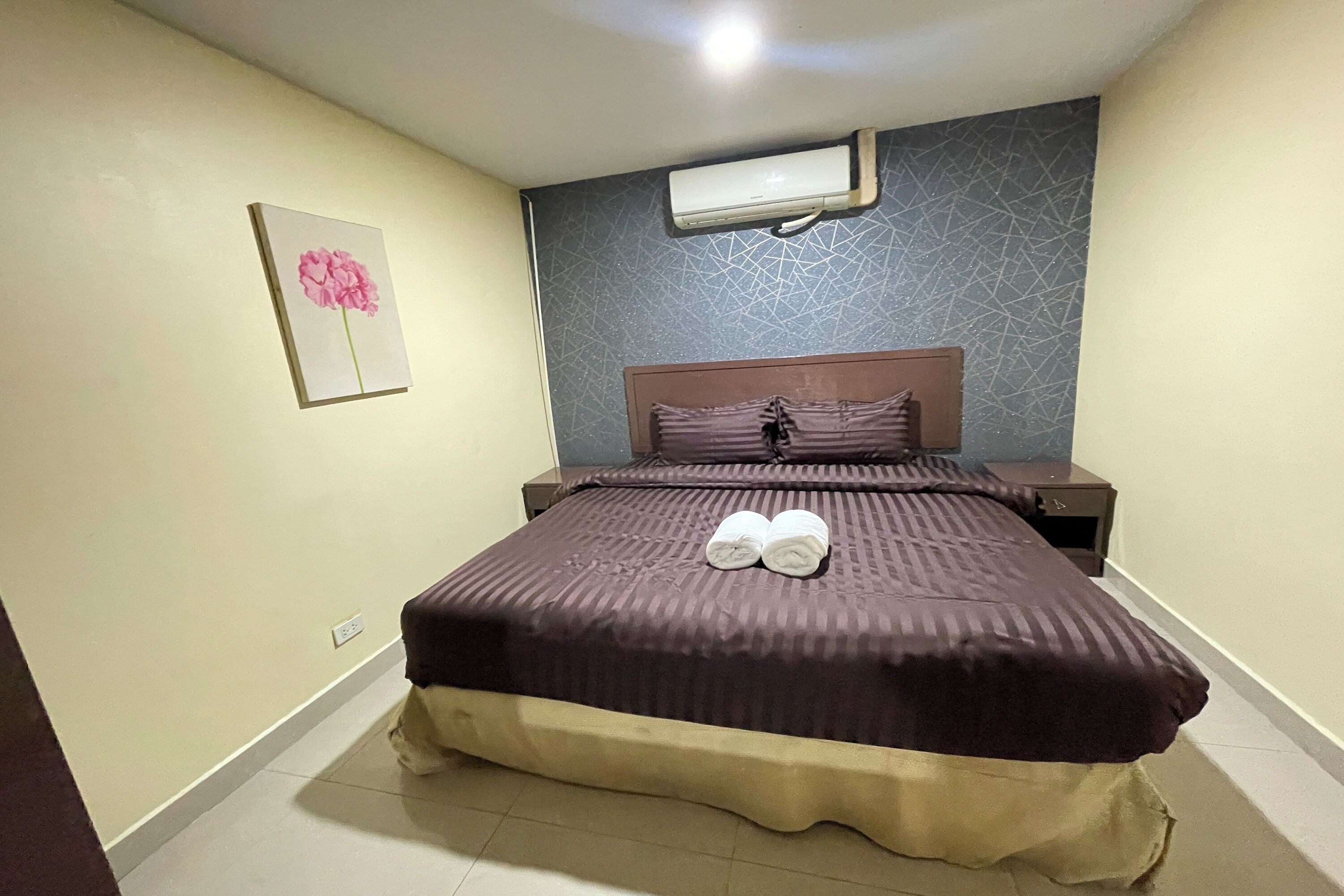 A neatly made bed with a brown cover, a wall-mounted air conditioner, and a framed picture of a flower on the wall.