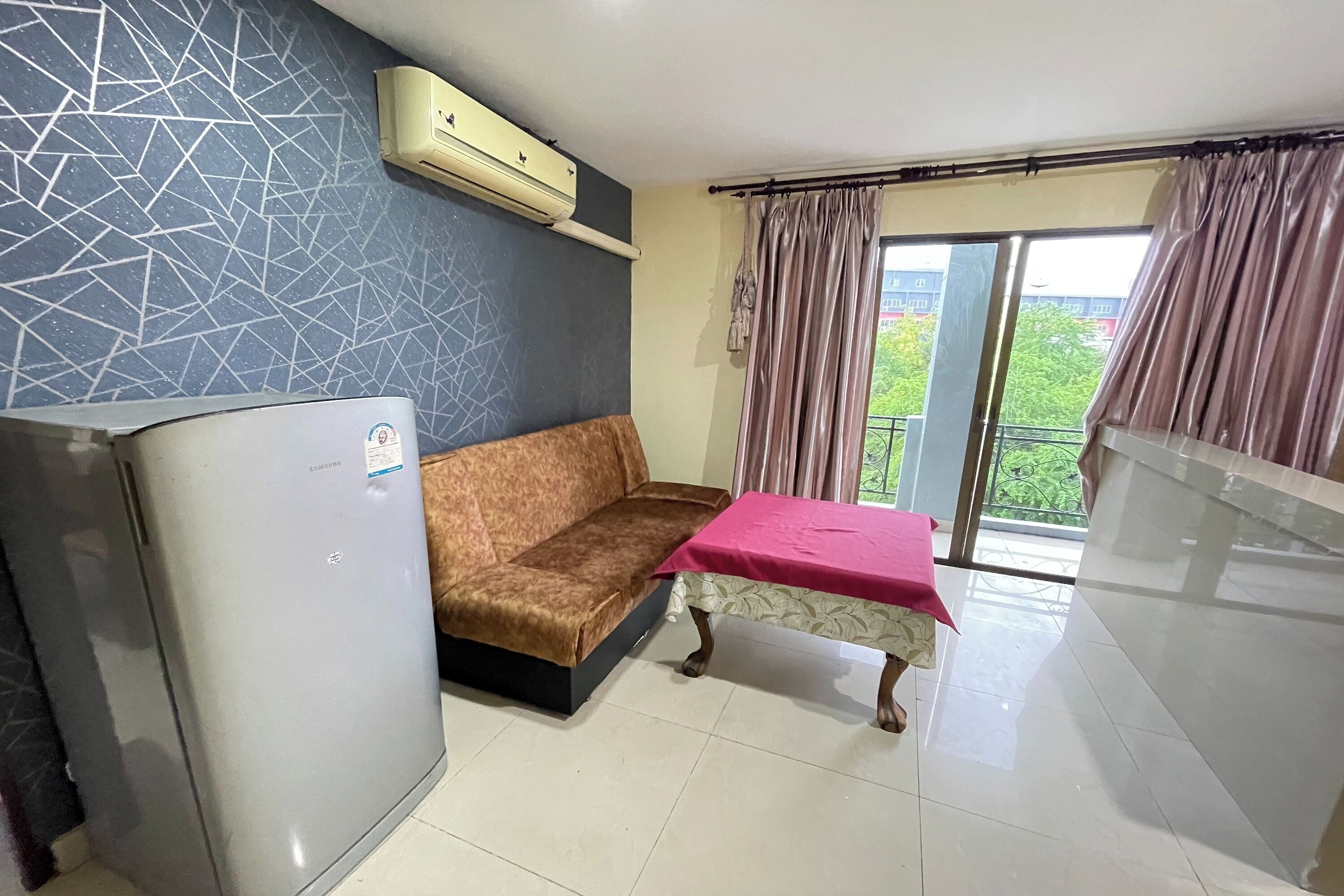 A room with a sofa, a table with a pink tablecloth, a refrigerator, and a window with curtains.