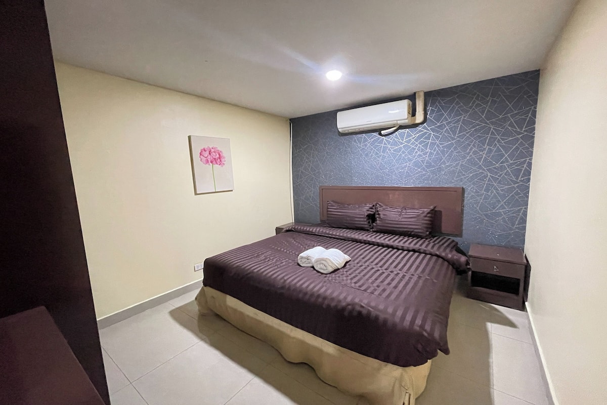 A hotel room with a bed, a nightstand, a wall-mounted air conditioner, and a picture of a flower on the wall.