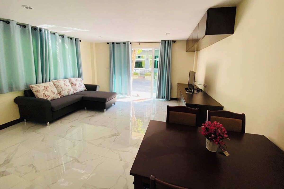 A modern living room with a sectional sofa, a dining table with chairs, and a flat-screen TV mounted on the wall.