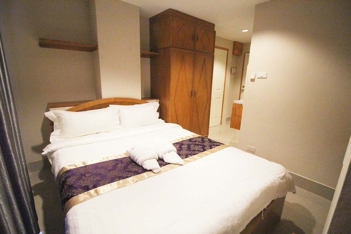 Ein ordentlich bezogenes Bett mit weißen Textilien und einem violetten Akzent, ein Holzschrank sowie ein Badezimmer mit weißem Waschbecken und Spiegel.
