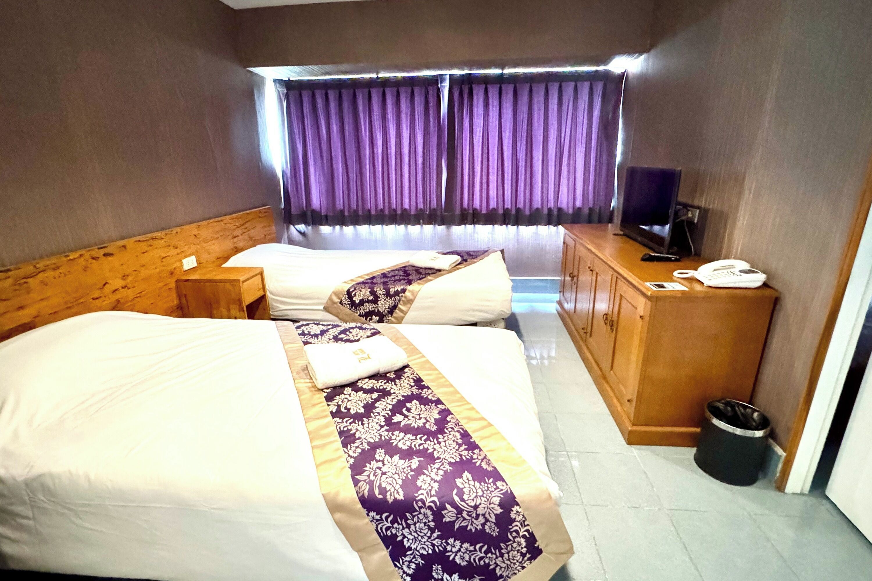 A hotel room with two beds, a wooden headboard, a desk with a telephone, and a window with purple curtains.