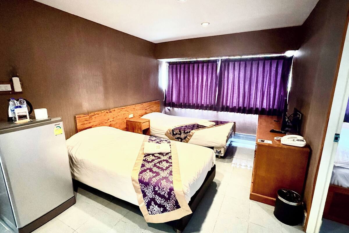 Ein Hotelzimmer mit zwei Betten, einem Kühlschrank, einem Nachttisch, einem Telefon und einem Fenster mit violetten Vorhängen.
